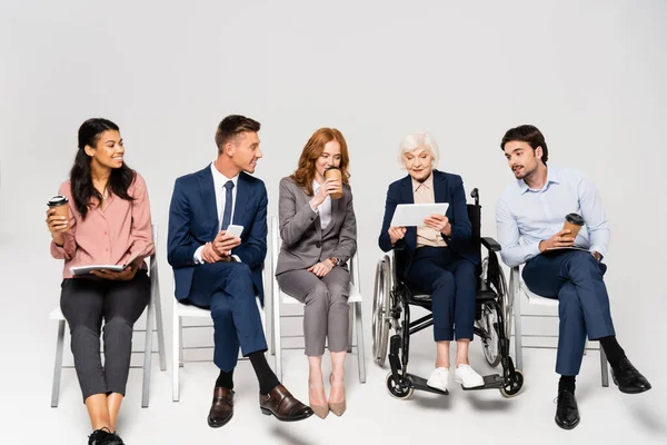 Empresário multiétnico sorridente com café para ir e pastas de papel sentado perto de empresária idosa em cadeira de rodas em fundo cinza — Fotografia de Stock