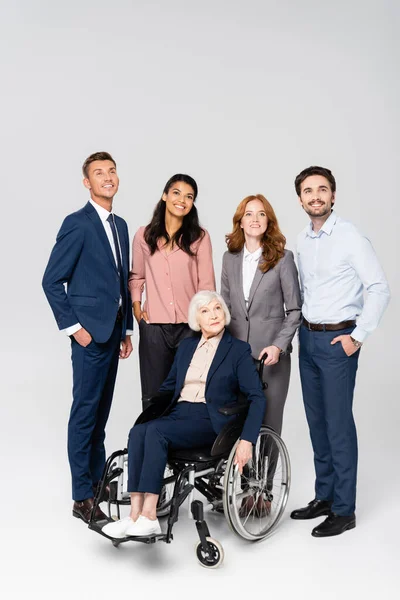 Smiling multiethnic businesspeople looking away near senior woman in wheelchair on grey background — Stock Photo