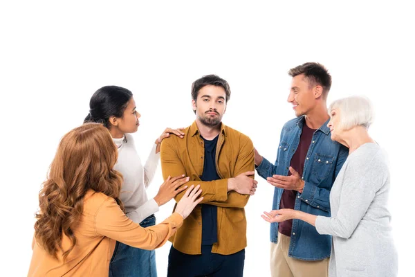 Triste hombre con los brazos cruzados de pie cerca de amigos multiétnicos aislados en blanco - foto de stock