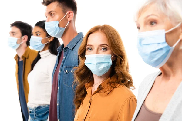 Mujer con máscara médica mirando a la cámara mientras está de pie cerca de personas multiétnicas sobre un fondo borroso aislado en blanco - foto de stock