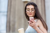 portrét mladé ženy v brýlích s kávou jít pomocí smartphone na ulici