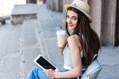 side view of young smiling woman with tablet and coffee to go sitting on steps clipart