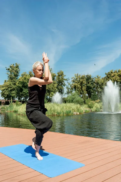 woman practicing yoga and standing on one leg on yoga mat near river in park