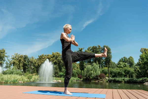 side view of woman practicing yoga on yoga mat and making namaste gesture near river in park