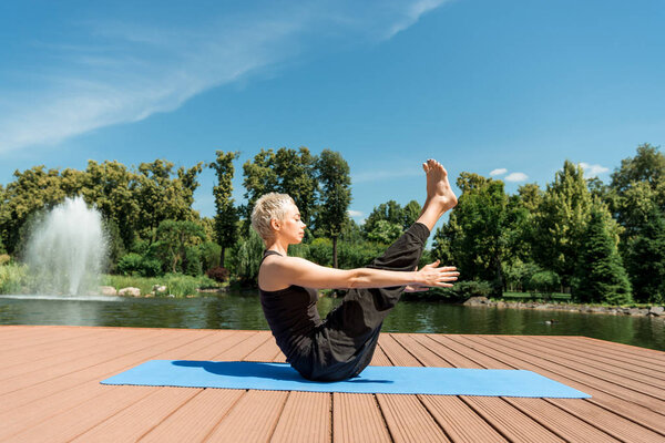 side view of athletic woman practicing yoga on yoga mat near river in park