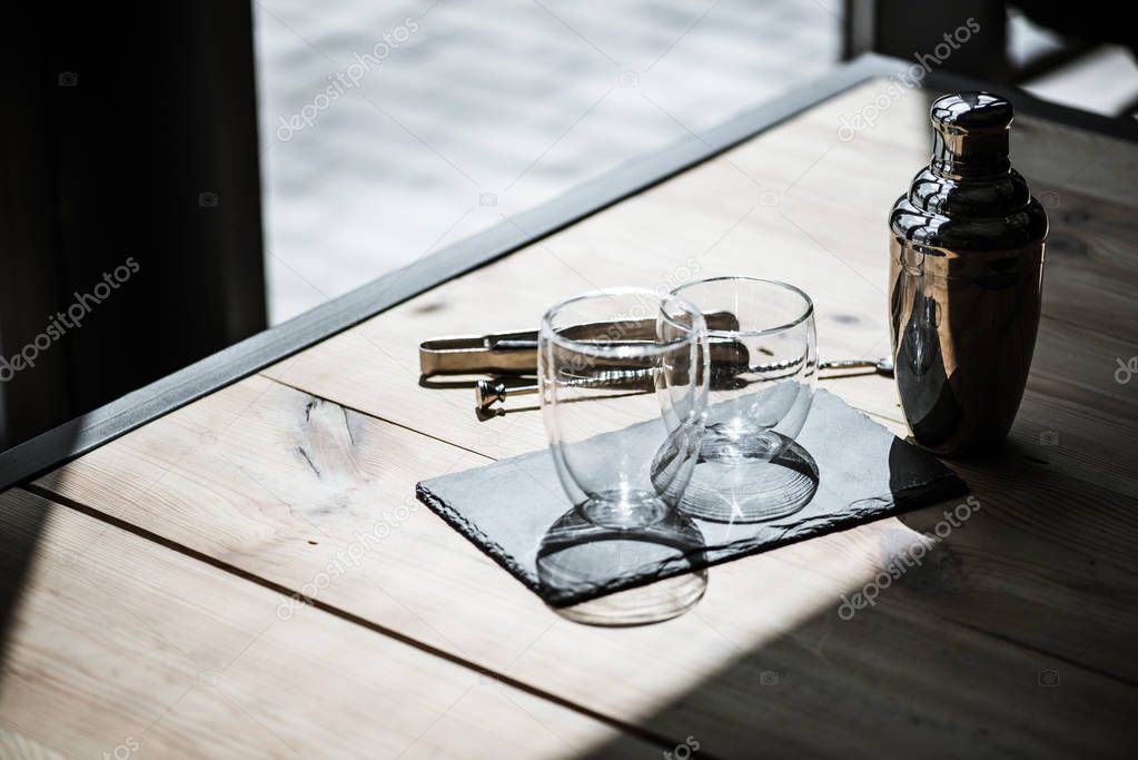high angle view of shiny empty glasses on slate board, shaker and tongs on wooden table 