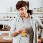 Χαρούμενος νεαρός άνδρας στέκεται με χυμό πορτοκαλιού στην κουζίνα στο σπίτι