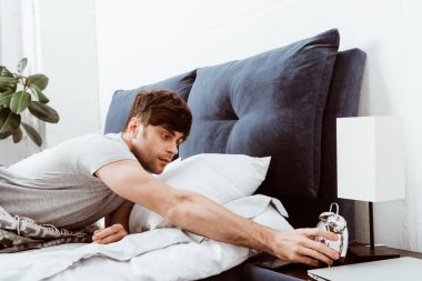 selective focus of young man turning off alarm clock in bed at home clipart