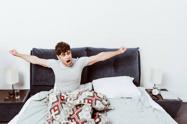handsome young man yawning and stretching in bed during morning time at home
