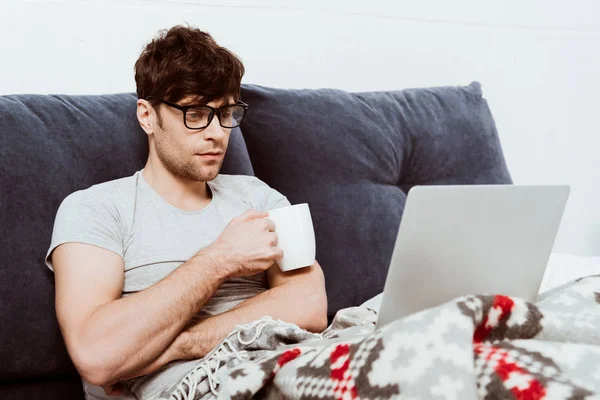 Focused Male Freelancer Eyeglasses Holding Coffee Cup Sitting Bed Laptop — Stock Photo, Image