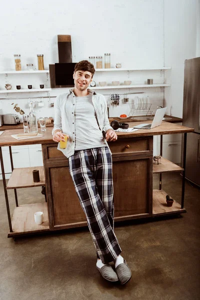 happy male freelancer with orange juice standing near table with laptop in kitchen