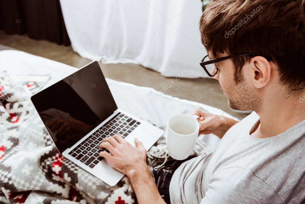 side view of male freelancer with coffee cup working on laptop with blank screen in bed at home