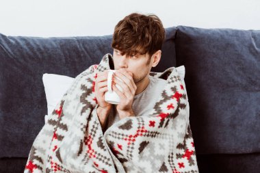 selective focus of sick man wrapped in blanket drinking tea in bed at home  clipart