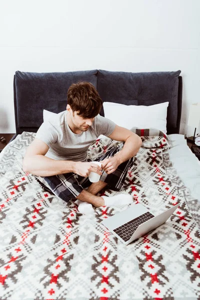 年轻的男性自由职业者与咖啡杯坐在床上与笔记本电脑在家里 — 免费的图库照片