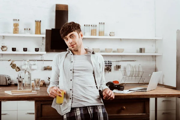 male freelancer with orange juice standing near table with laptop in kitchen at home