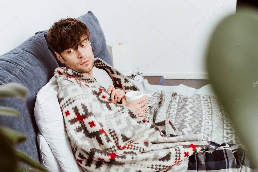 selective focus of sick man wrapped in blanket sitting with cup of tea in bed at home 
