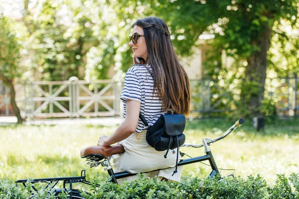 Visão traseira da jovem mulher em óculos de sol sentado na bicicleta retro e falando no smartphone no parque — Fotografia de Stock