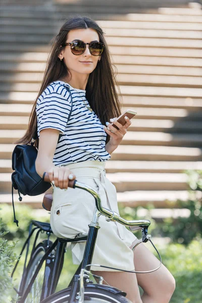 Вид сбоку привлекательной улыбающейся женщины со смартфоном, опирающейся на велосипед на улице — стоковое фото