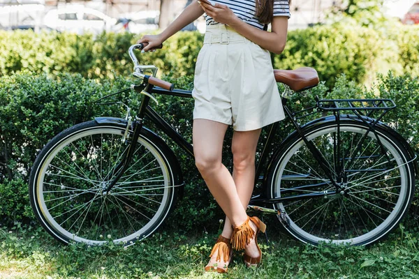 Cropped shot of woman with smartphone and retro bicycle standing in park — Stock Photo