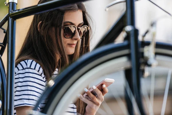 Portrait of young woman using smartphone while sitting near retro bicycle on street — Stock Photo