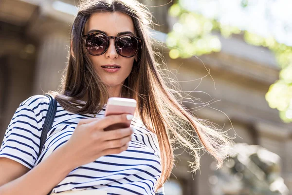 Low angle view of young attractive woman in sunglasses using smartphone on street — Stock Photo