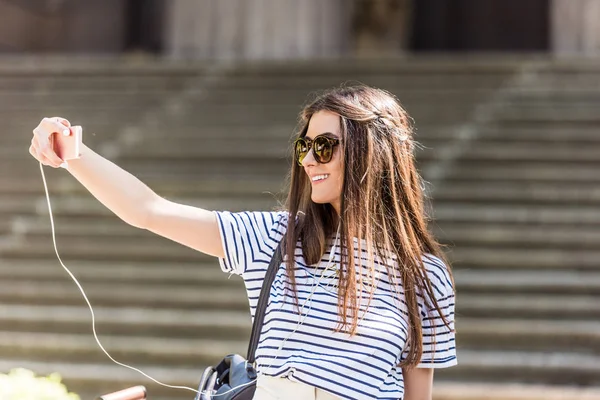 Retrato de mujer sonriente atractiva en auriculares tomando selfie en teléfono inteligente en la calle - foto de stock