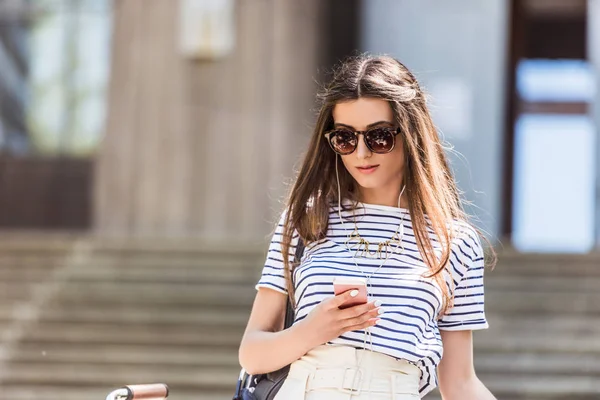 Ritratto di giovane donna attraente in auricolare utilizzando smartphone sulla strada — Foto stock