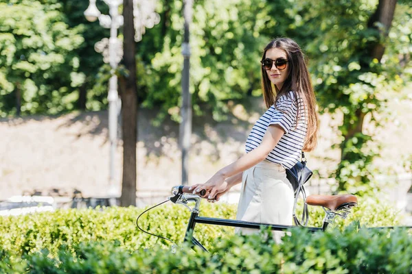 Vista laterale di elegante giovane donna in occhiali da sole con bicicletta retrò nel parco — Foto stock