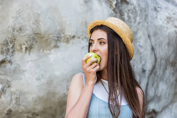 Portrait of young stylish woman in hat eating fresh apple on street — Stock Photo