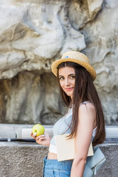 Vista lateral de la joven mujer sonriente en sombrero con manzana fresca en la calle - foto de stock