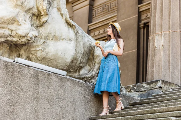 Giovane donna elegante in cappello con mela fresca in piedi su gradini sulla strada — Foto stock