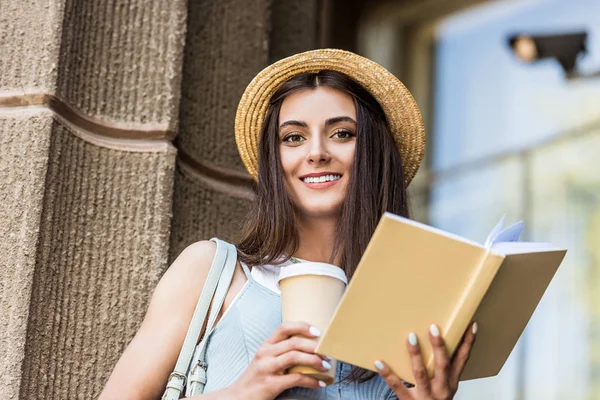 Jolie femme souriante avec livre ouvert et café pour aller dans la rue — Photo de stock