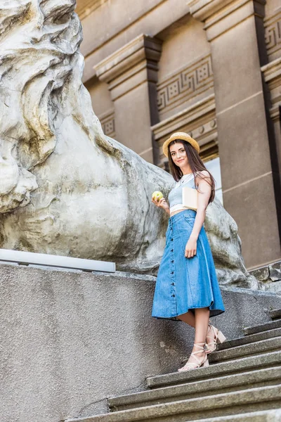 Young stylish woman in hat with fresh apple standing on steps on street — Stock Photo