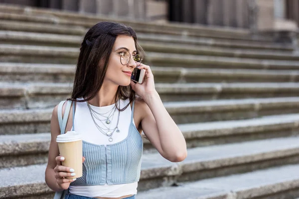 Retrato de mujer joven con café para ir a hablar en smartphone en la calle - foto de stock