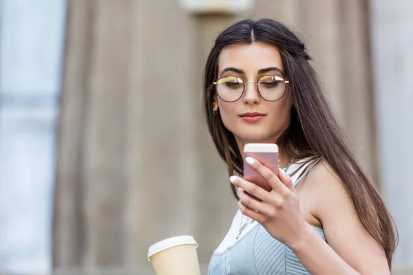 Portrait of young woman in eyeglasses with coffee to go using smartphone on street — Stock Photo