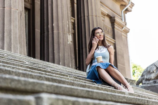 Jovem mulher sorridente com café para ir falar no smartphone enquanto sentado em passos na rua — Fotografia de Stock