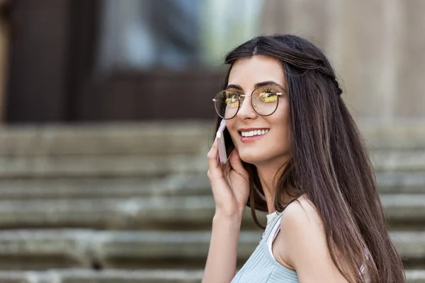 Joven mujer sonriente en gafas que habla en el teléfono inteligente en la calle - foto de stock