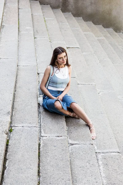 Jovem mulher elegante em óculos e saia jeans descansando em passos na rua — Fotografia de Stock