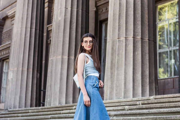Молодая стильная женщина в очках и джинсовой юбке стоя на ступеньках — стоковое фото