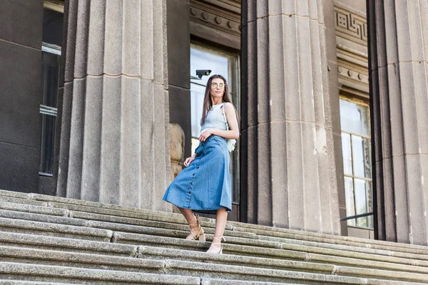 Young stylish woman in eyeglasses and denim skirt standing on steps — Stock Photo