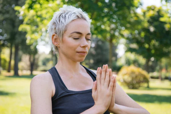 Donna che pratica yoga con gli occhi chiusi e fare gesto namaste con le mani nel parco — Foto stock