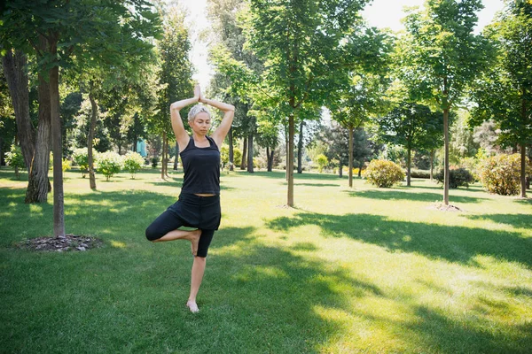Женщина практикует йогу в позе дерева (Vrksasana) на траве в парке — стоковое фото