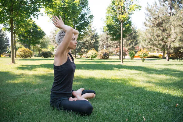 Vista lateral de la mujer practicando yoga en pose de loto con las manos levantadas haciendo gesto namaste sobre hierba en parque - foto de stock