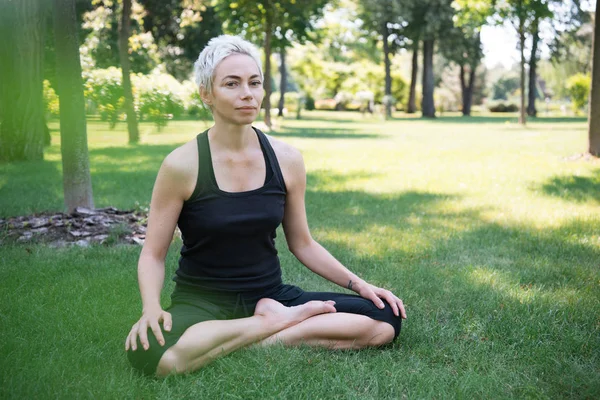 Mujer practicando yoga en pose de loto y mirando cámara sobre hierba en parque - foto de stock