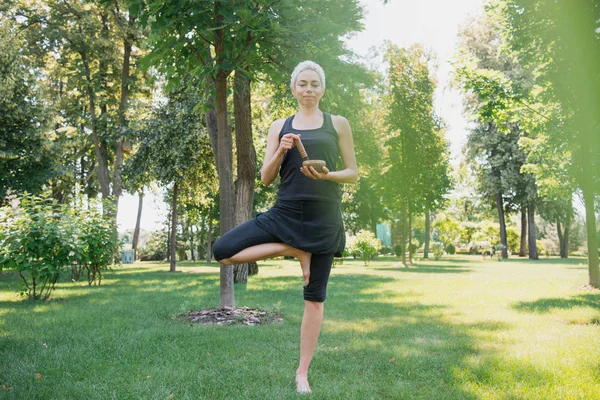 Atractiva mujer practicando yoga en pose de árbol y haciendo sonido con cuenco de canto tibetano en parque - foto de stock