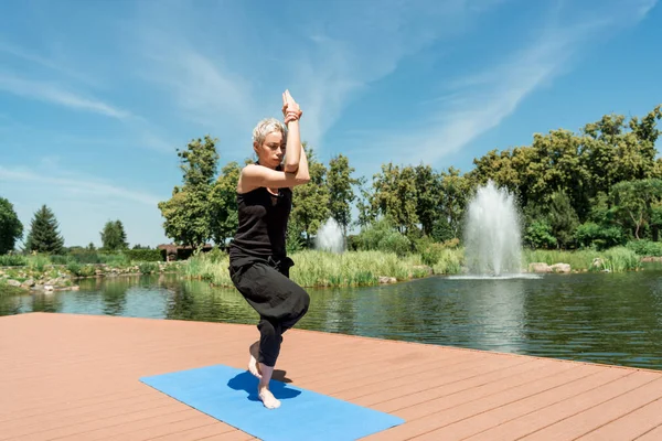 Athletic woman practicing yoga and standing on one leg on yoga mat near river in park — Stock Photo