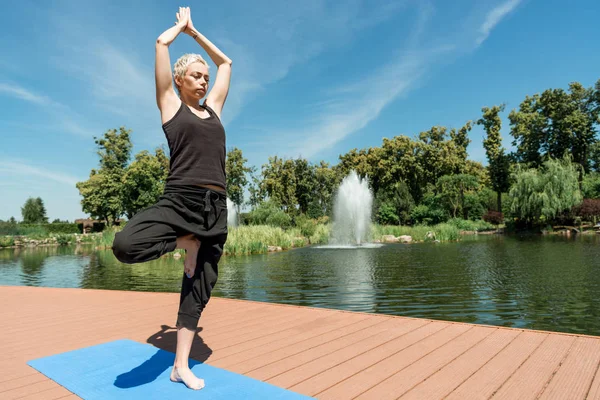 Athletische Frau praktiziert Yoga in Baumpose (vrksasana) auf Yogamatte in Flussnähe im Park — Stockfoto