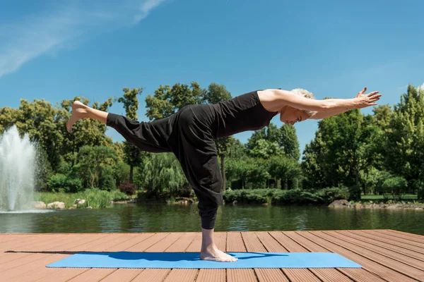 Seitenansicht einer Frau, die Yoga im Krieger iii (virabhadrasana iii) praktiziert, posiert auf einer Yogamatte in der Nähe des Flusses im Park — Stockfoto