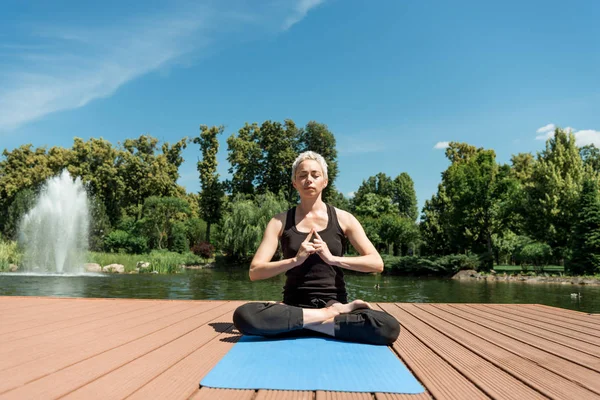 Athletic woman practicing yoga in lotus pose on yoga mat near river in park — Stock Photo