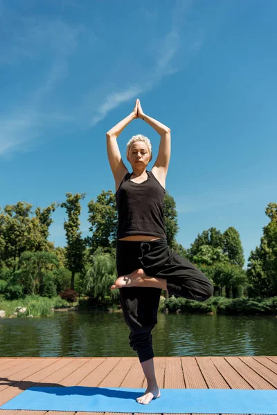 Mulher praticando ioga em pose de árvore (Vrksasana) e fazendo gesto namaste no tapete de ioga perto do rio no parque — Fotografia de Stock